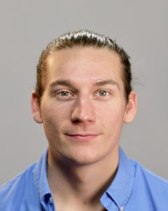 A headshot photograph of Oliver Robertson staring into the camera. He has brown hair tied back and is wearing a blue shirt. 