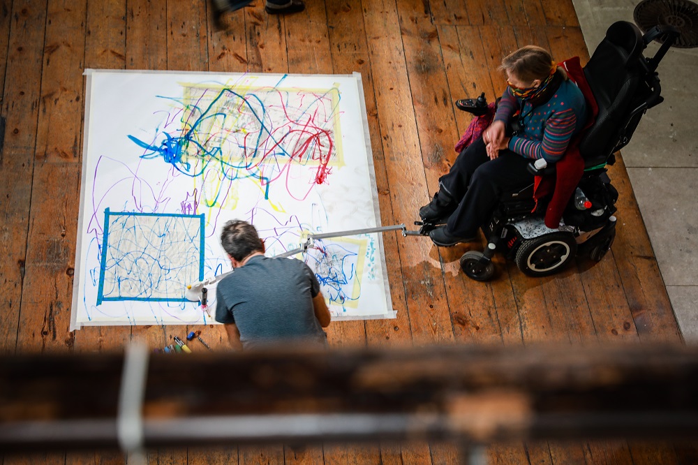 An ariel view of a man and a woman who is a wheelchair user looking at a painting they made.
