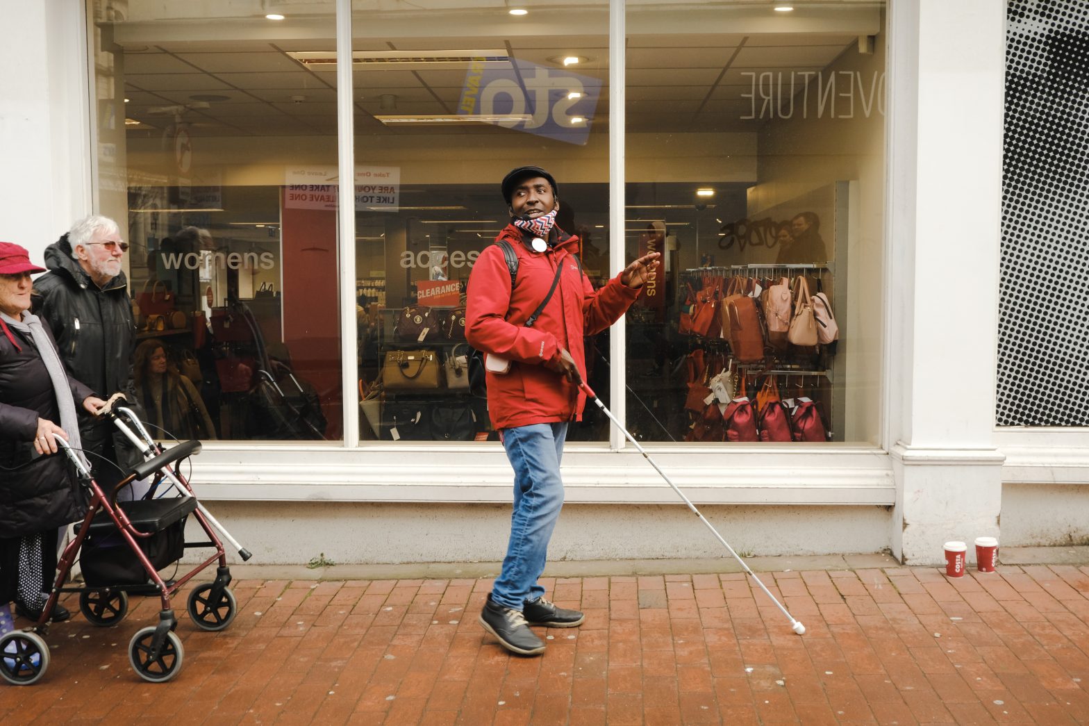 A man is walking past a shop using his cane. He is wearing blue jeans and a bright red jacket.