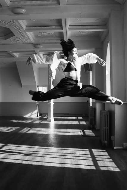 A black and white image of a dancer who is leaping through the air in a dance studio.