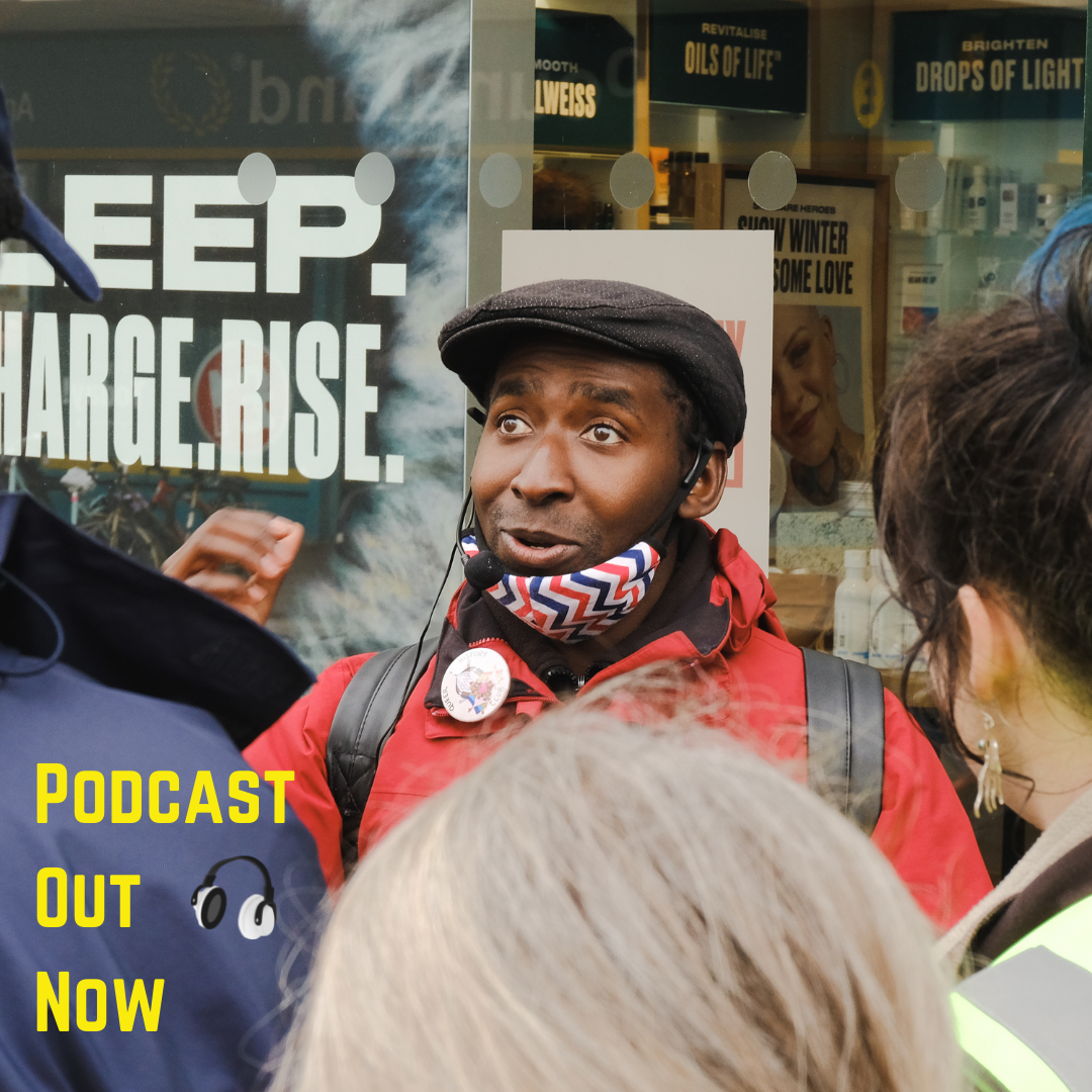 Photo of a man speaking to a group of people outside of a shop. Text Reads: 'Podcast Out Now' in yellow.