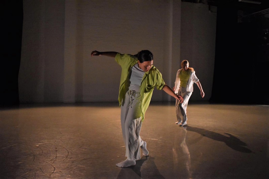 Two women dance in a studio. They are wearing white and greens. They are mid step and have their arms open.
