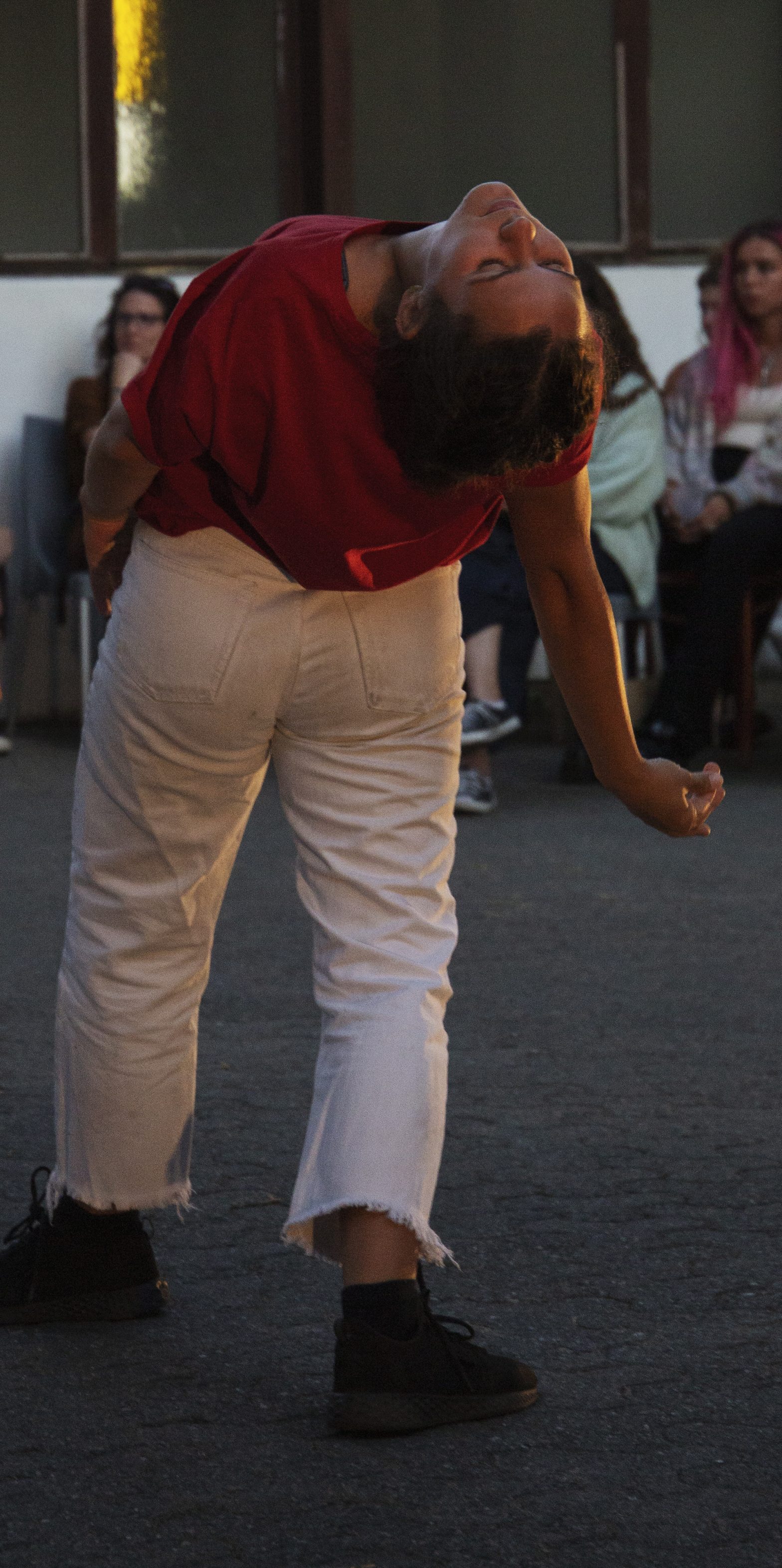 A woman is dancing. She is wearing white trousers and a red top and leans backwards.