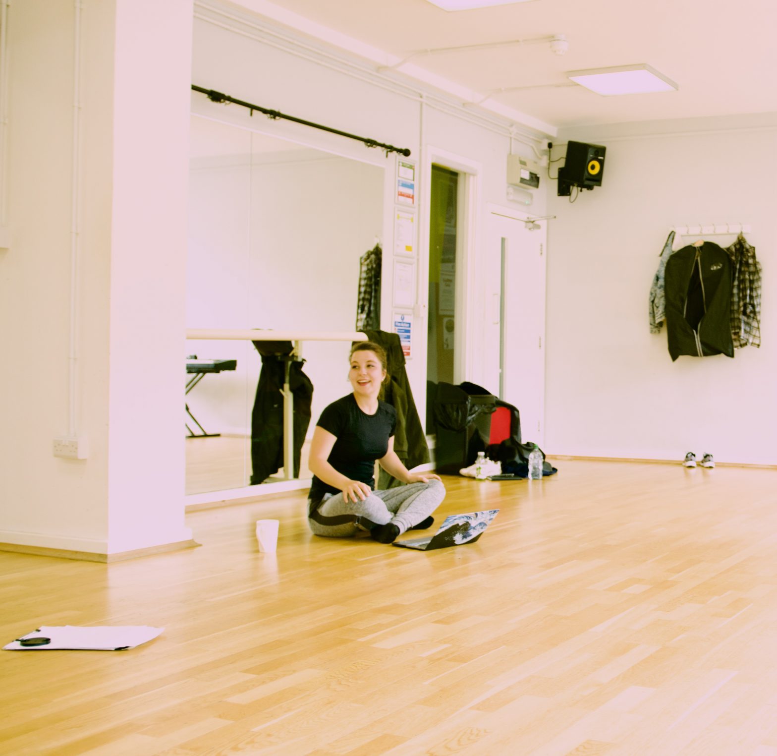 A woman is sat in a dance studio. She has her hair back is is wearing a dark top and grey trousers.