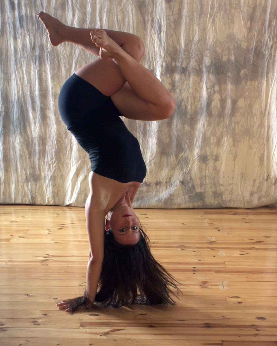 A woman is doing a hand stand with her legs twisted in. She is wearing black and her long hair is down.