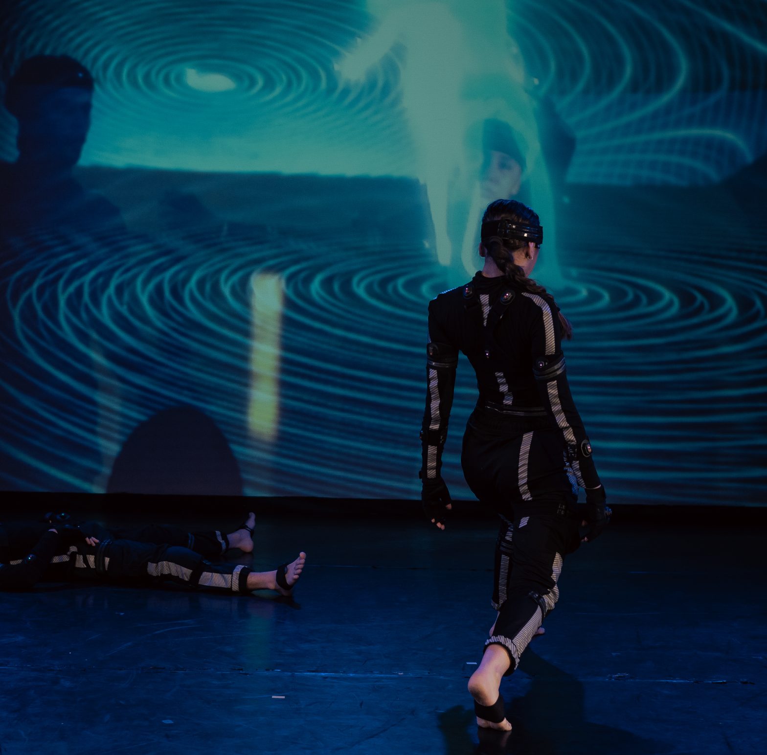 One dancer lies on the floor as the other walks towards the back on the theatre. Both wear motion capture, their avatars projected behind them.