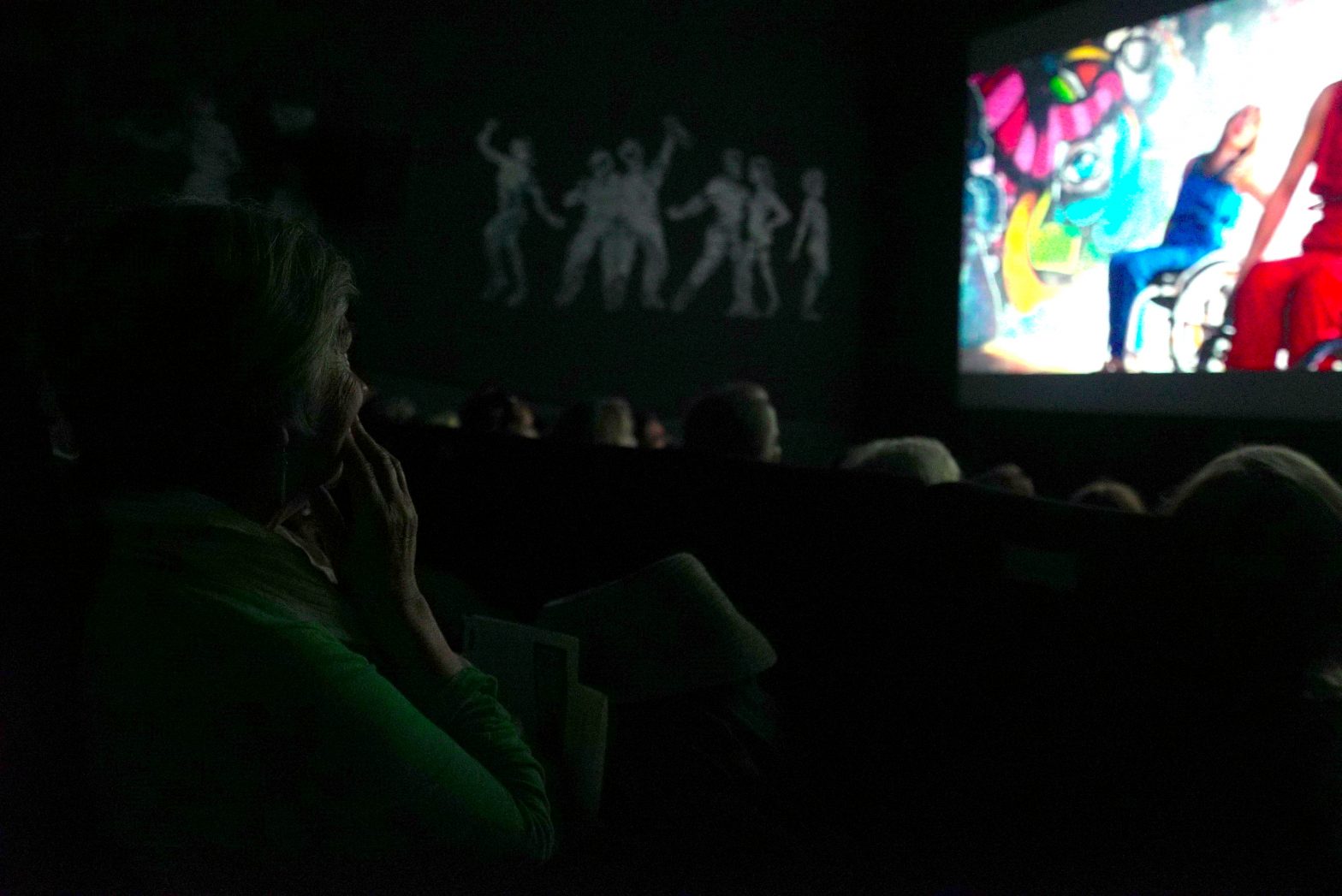 A film is showing on the cinema showing two dancers in bright outfits who use wheelchairs. The camera is focused on a woman watching the film, her hand to her mouth.
