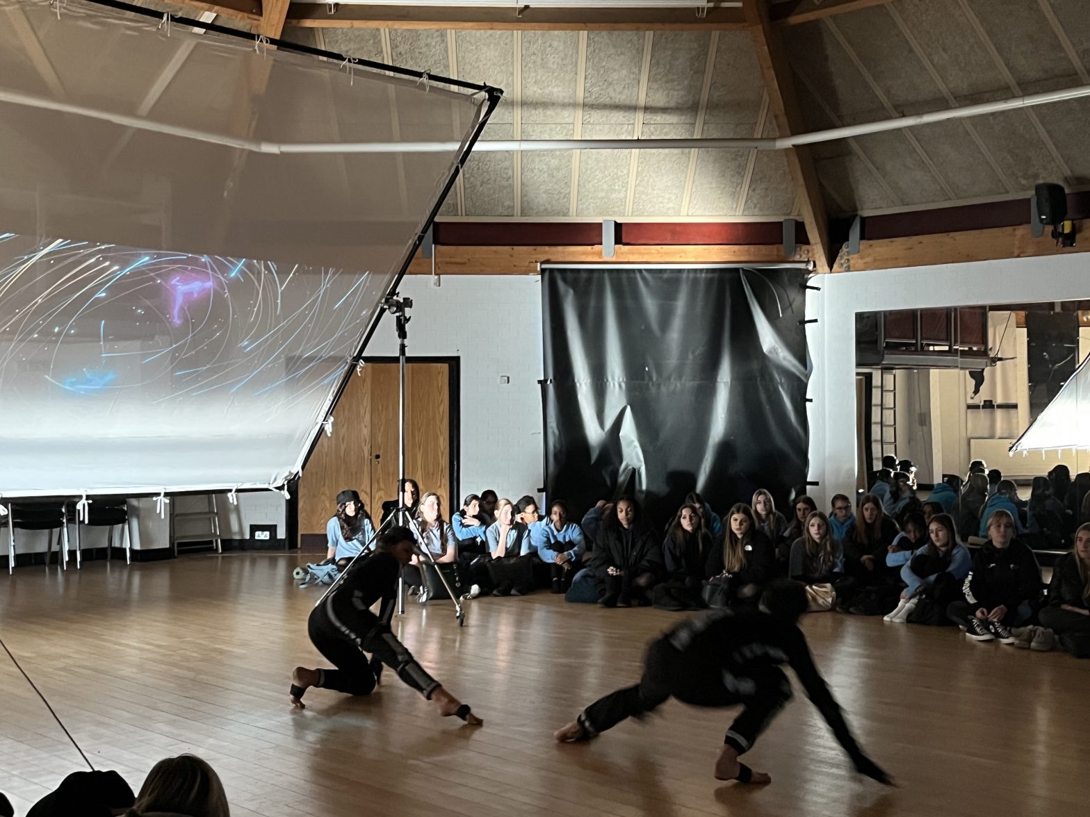 School children sit watching a motion capture dance performance with a large screen.