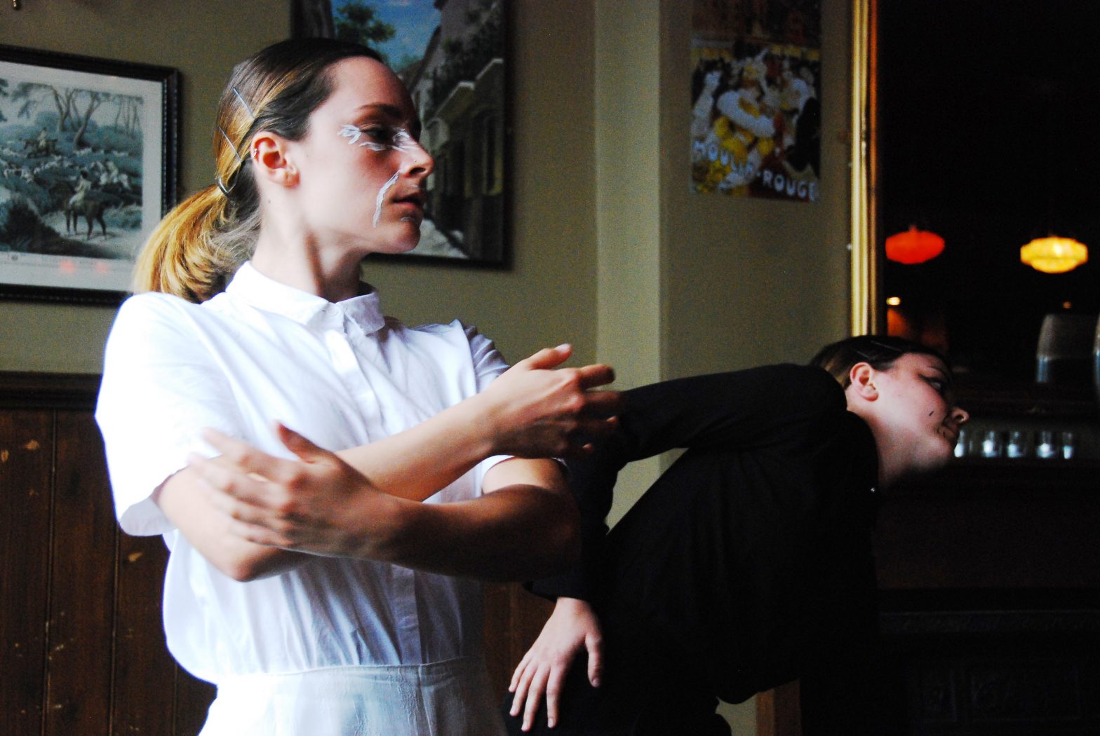 Two women are dancing in a pub function room. One is wearing white, the other black. One is stood up with her arms crossed, the other leaning to the side.