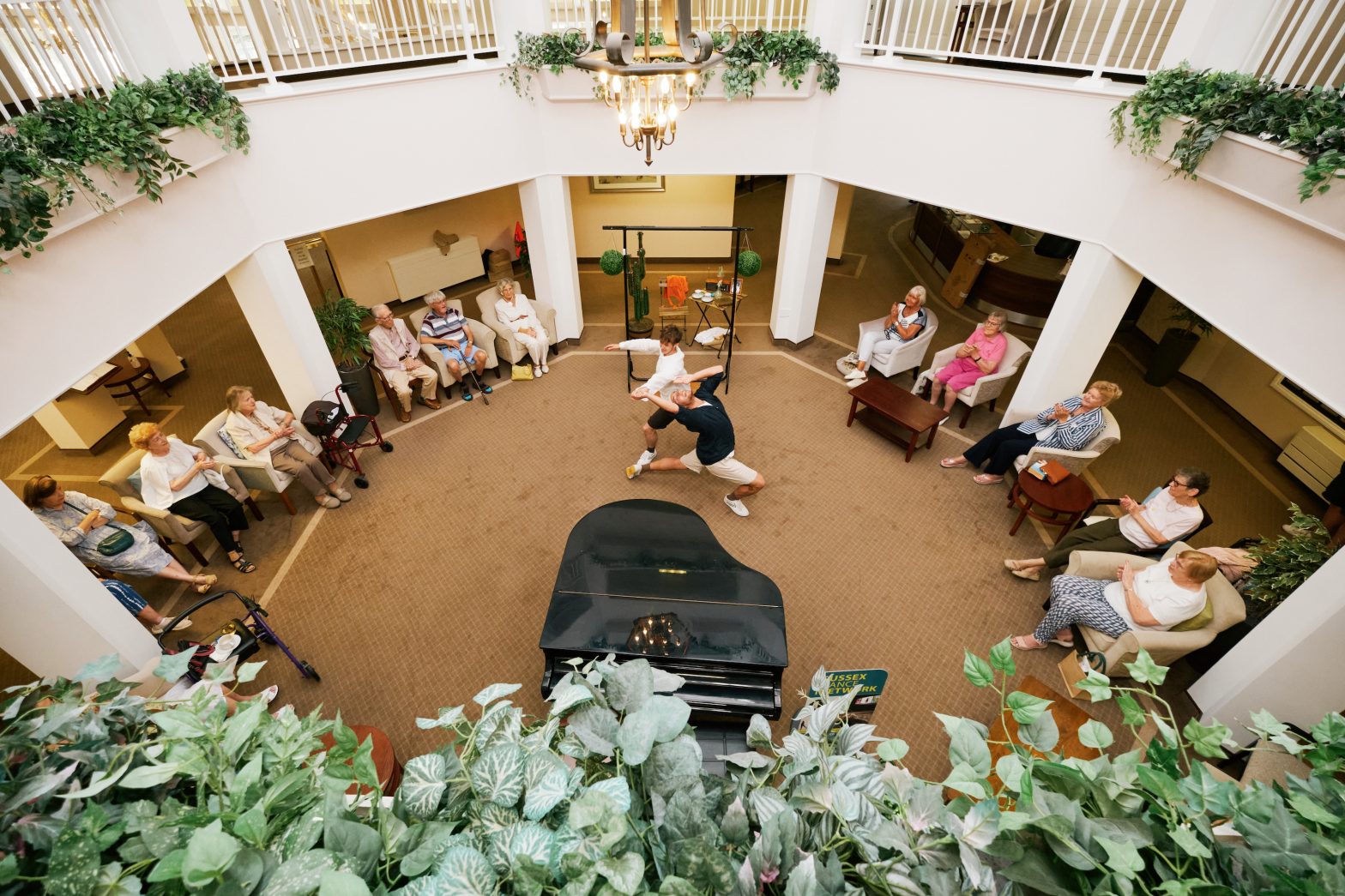 Two male dancers are performing in the centre of a retirement village. The view is from a balcony and look down on the dancers and audience. The audience are sat around the edge of the space. There is also a piano, plants and a carpeted floor.