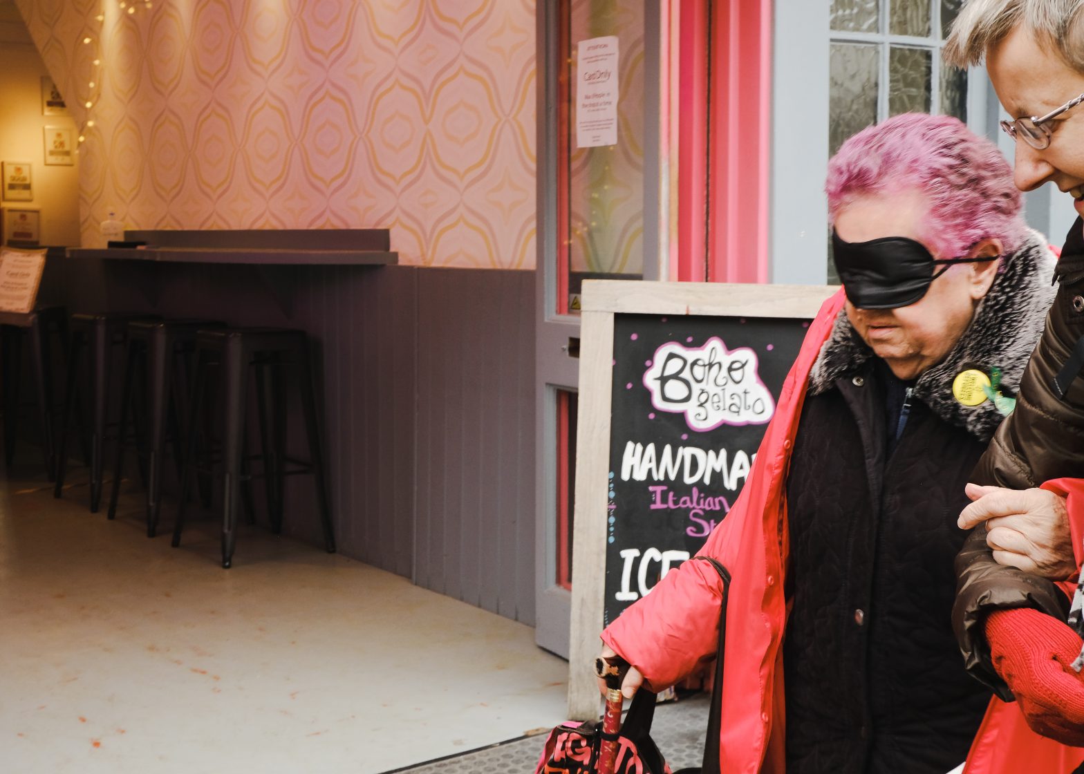 A woman with pink hair is wearing a blindfold. She is being guided by another woman outside an ice cream shop.