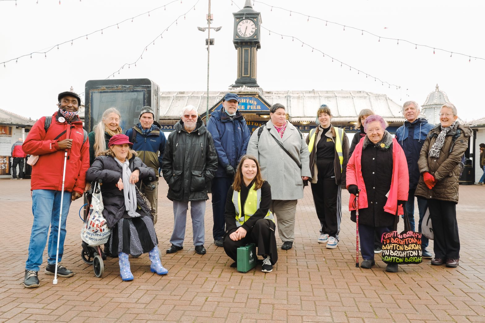 A group of people pose for a photo outside Brighton Pier. SDN team members are wearing high visibility vests. The rest of the group are an older group of the community with experiences of visual impairment and dance artist Ebony Rose Dark.